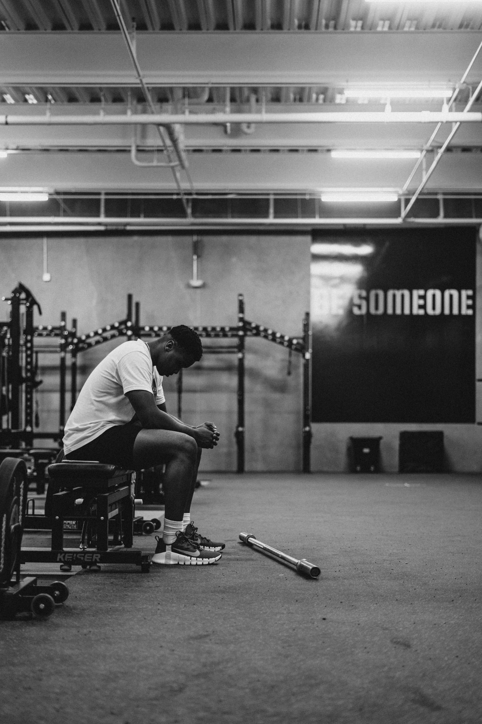 Sportiv așezat, epuizat după un antrenament intens, lângă inscripția motivațională „Be Someone” pe peretele sălii